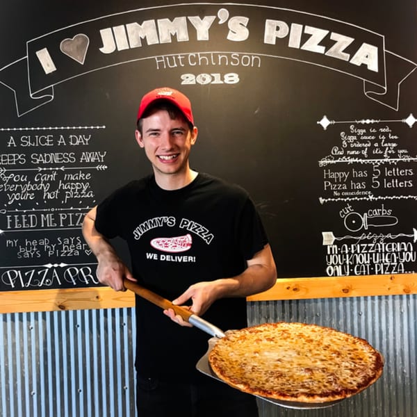 A smiling Jimmy's Pizza employee holds a freshly baked cheese pizza on a peel in front of a chalkboard.