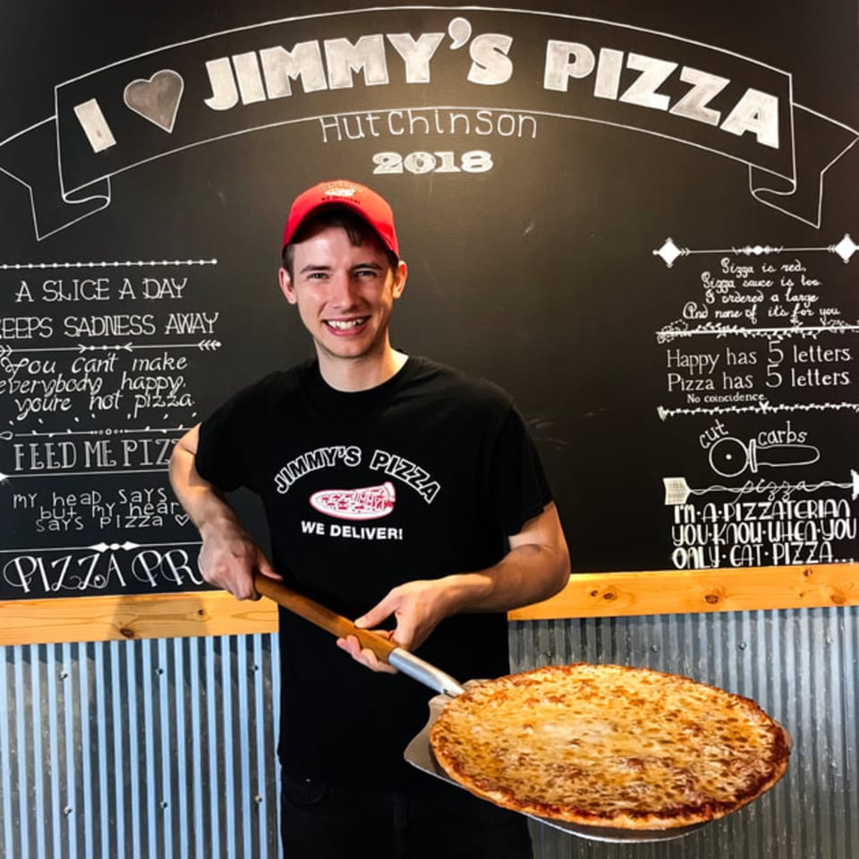 A smiling Jimmy's Pizza employee holds a freshly baked cheese pizza on a peel in front of a chalkboard.