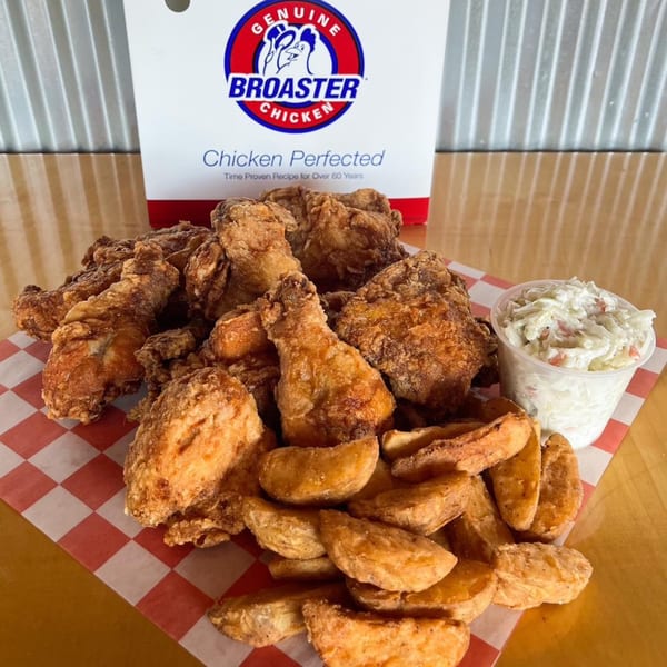 A pile of crispy fried chicken, potato wedges, and coleslaw on checkered paper with a Broaster Chicken box.