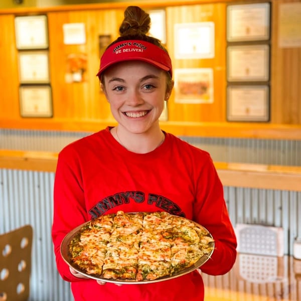 Smiling Jimmy's Pizza employee in red uniform holding a freshly baked pesto chicken pizza.