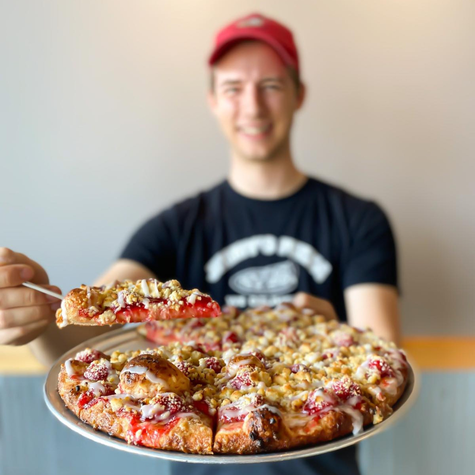 A smiling Jimmy's Pizza employee holding a dessert pizza with strawberry, crumble, and icing.