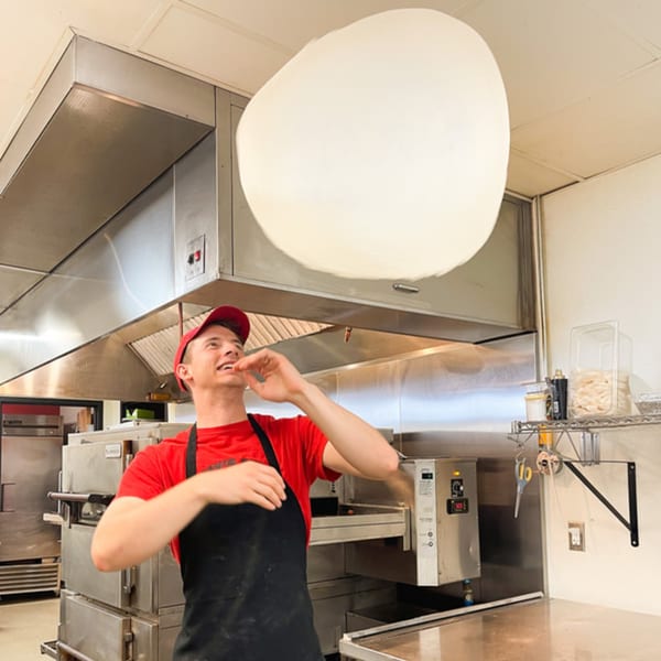 A Jimmy's Pizza chef in a red shirt and black apron expertly tosses pizza dough high in the air.