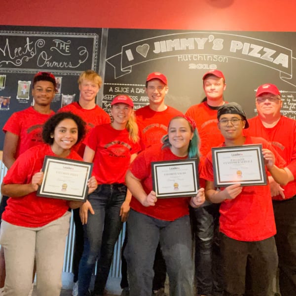 Nine smiling Jimmy's Pizza staff members in red shirts and hats, holding awards in front of a chalkboard.