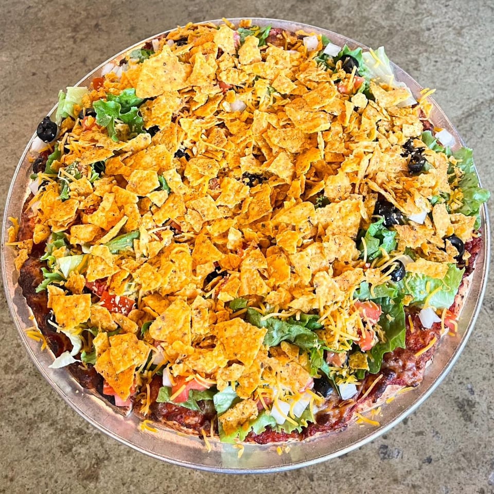 A taco pizza from Jimmy's Pizza, topped with lettuce, tomatoes, black olives, cheese, and crushed tortilla chips.