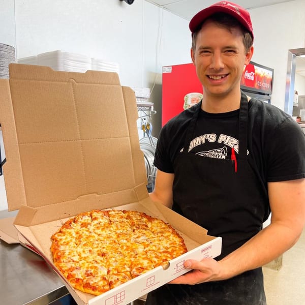 Smiling Jimmy's Pizza employee in a red hat and black apron holding an open pizza box with a cheese pizza.