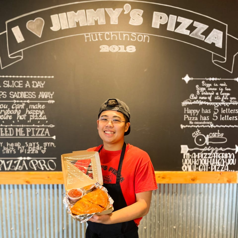 A smiling Jimmy's Pizza employee holds a calzone and sauce in a takeout box against a chalkboard wall.