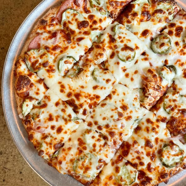 A close-up of a Jimmy's Pizza with melted cheese, ham, and sliced pickles on a metal pan.