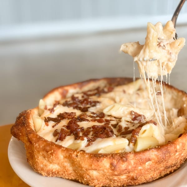 A deep-dish mac and cheese pizza with bacon, with a fork lifting a cheesy bite.