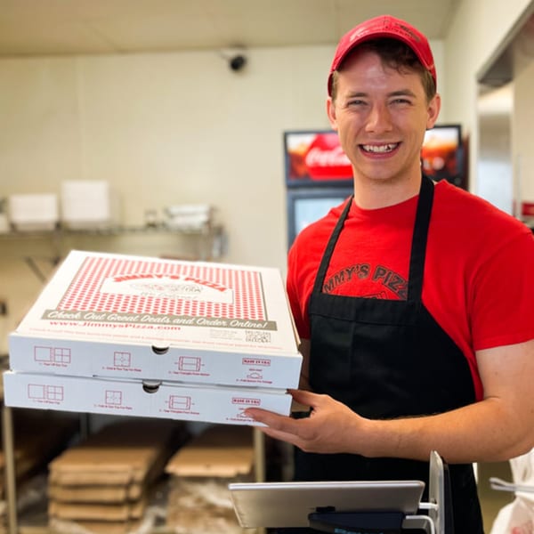 A smiling Jimmy's Pizza employee in a red shirt and apron holds two pizza boxes.