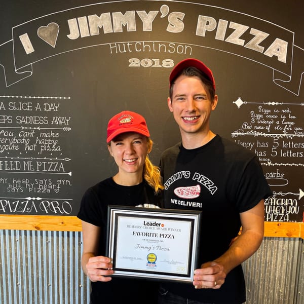 Two smiling Jimmy's Pizza employees hold a "Favorite Pizza" award in front of a chalkboard wall.