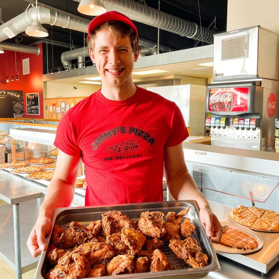 Smiling Jimmy's Pizza employee in a red shirt and cap holding a tray of fried chicken.