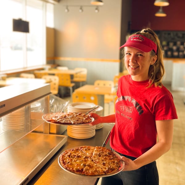 A smiling Jimmy's Pizza employee in a red visor and shirt holds two freshly baked pizzas.