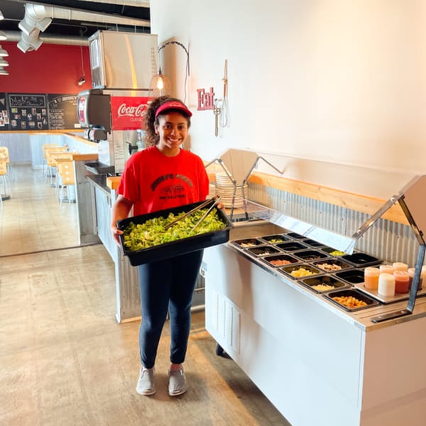 A smiling Jimmy's Pizza employee holds a tray of lettuce next to a salad bar with various toppings.