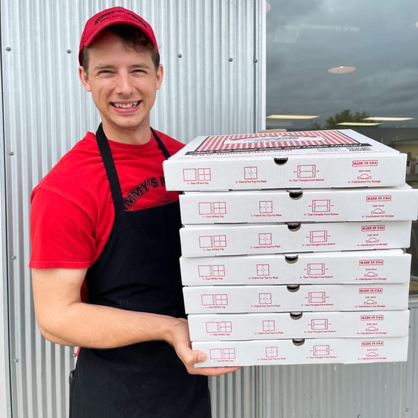 A smiling Jimmy's Pizza employee in a red shirt and cap holds a tall stack of pizza boxes.