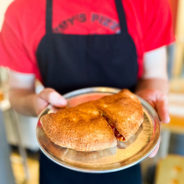 A person in a red shirt and black apron holds a metal plate with a golden-brown calzone.