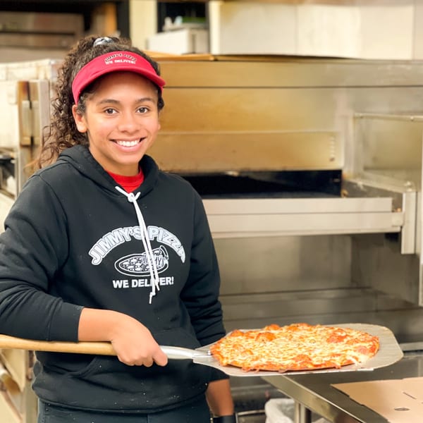 A smiling Jimmy's Pizza employee holding a freshly baked pepperoni pizza on a peel.