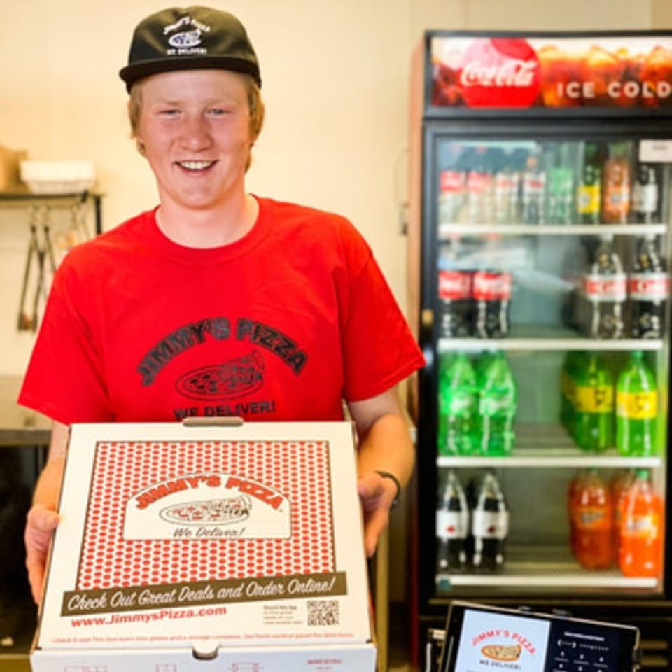 Smiling Jimmy's Pizza employee in a red shirt and hat holding a pizza box.